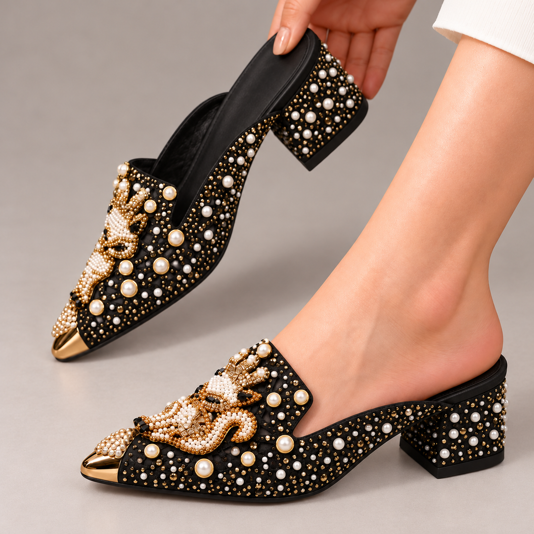 Black Odette mules shown worn on one foot with the matching mule held behind, highlighting pearl and gold embellishments and pointed gold toe caps.