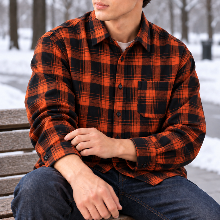 Model seated on a bench wearing an orange and black plaid flannel shirt, with snow in the background.